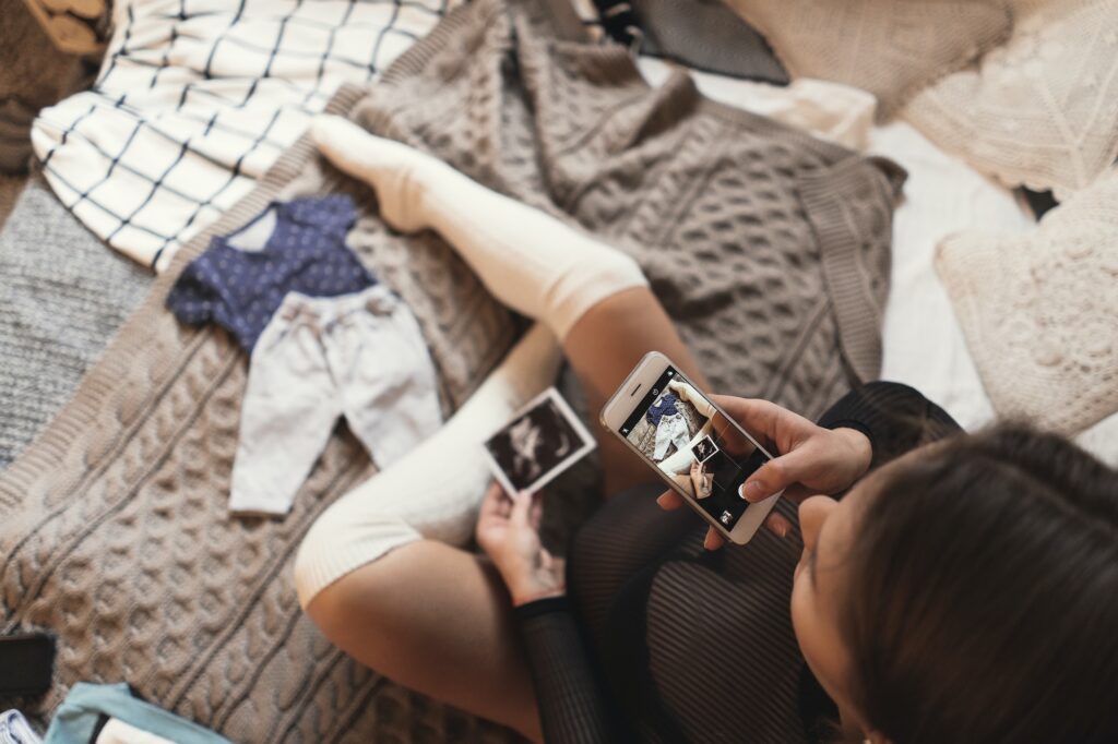 Pregnant young woman taking pictures of ultrasound photos with smartphone while relaxing on a bed