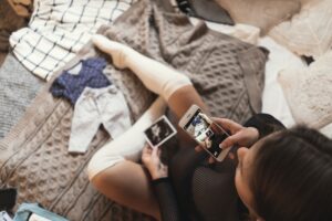 Pregnant young woman taking pictures of ultrasound photos with smartphone while relaxing on a bed